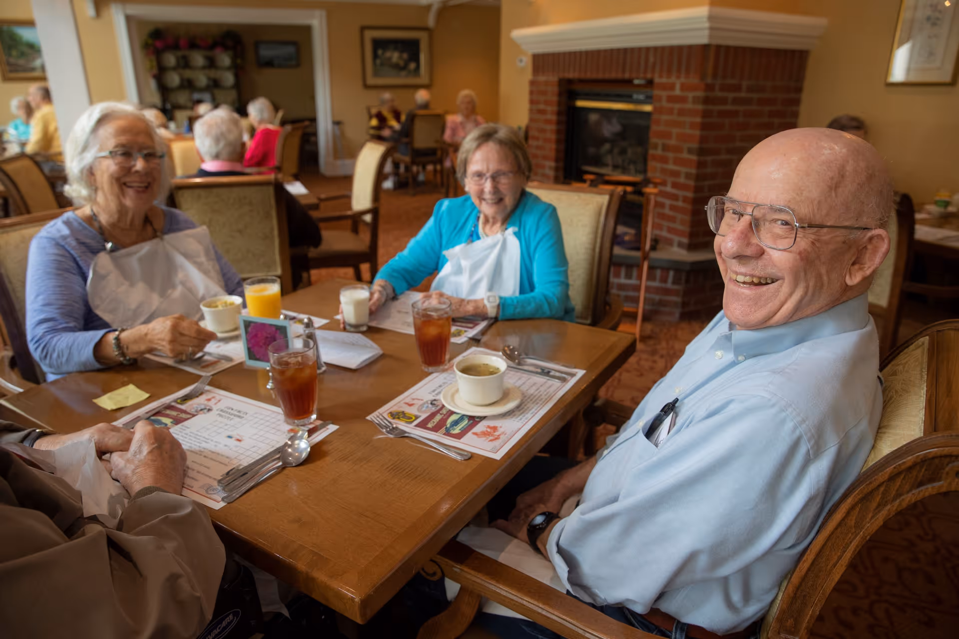 A group of elderly people sitting around a dining table in a senior living facility, smiling and enjoying drinks including iced tea, orange juice, and soup. The room has warm lighting, a brick fireplace, and other residents seated at tables in the background.