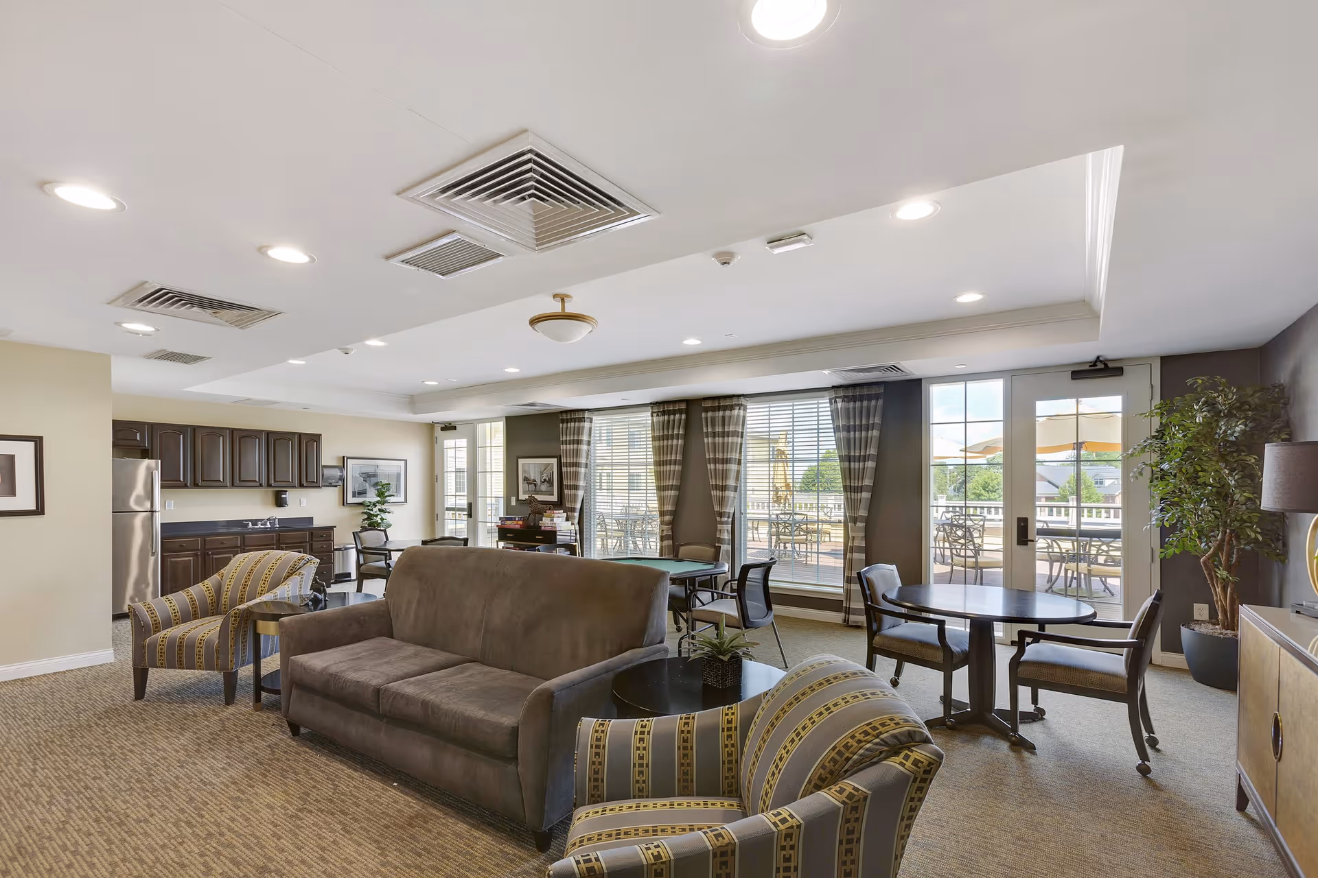 A spacious senior living common area with a brown sofa, two patterned armchairs, round tables with chairs, large windows with striped curtains, and a door leading to an outdoor patio with tables and umbrellas. The room also features a kitchenette with dark cabinets and a stainless steel refrigerator.