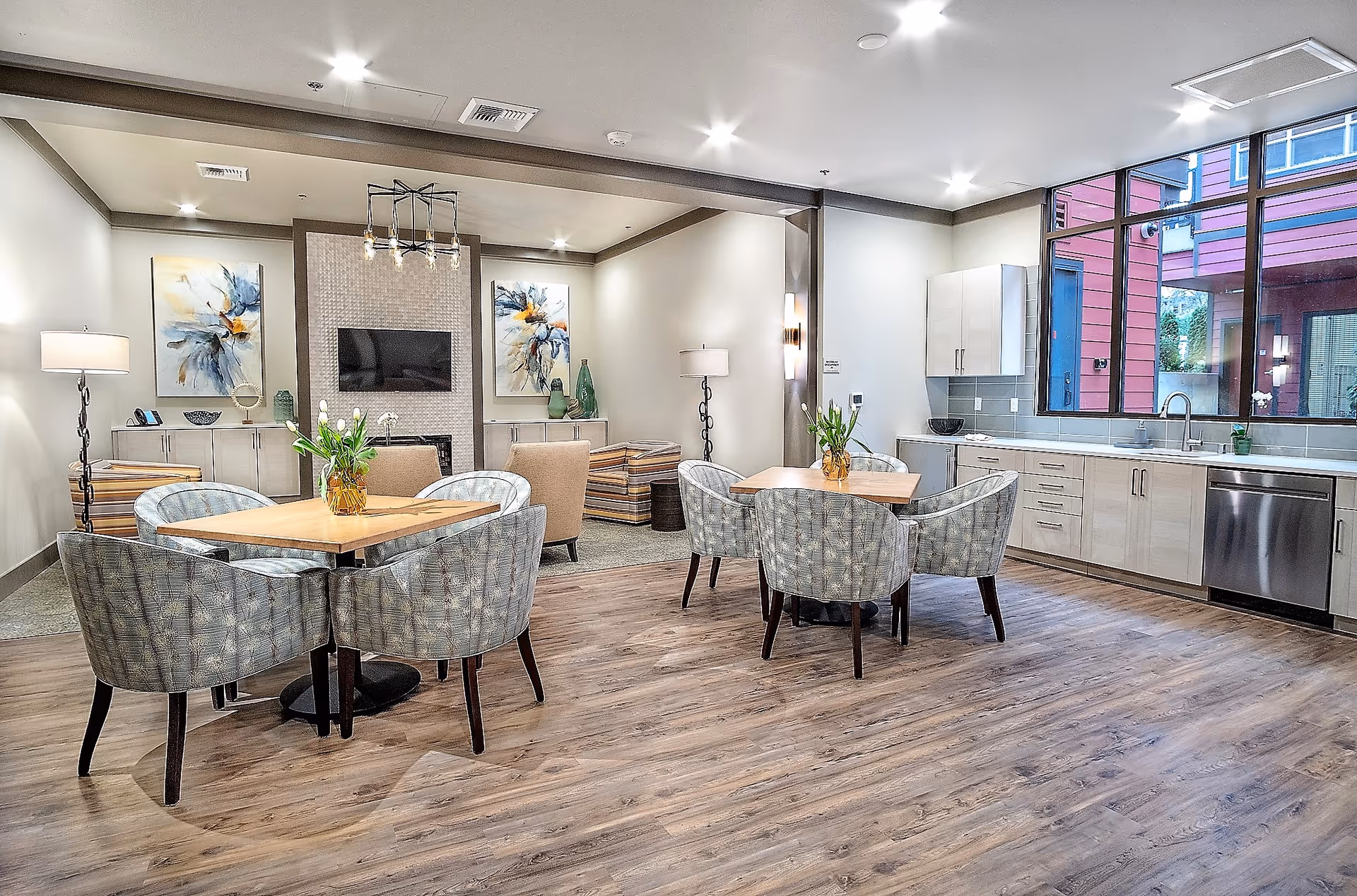 Bright communal dining and lounge area with round tables and upholstered chairs, a kitchenette on the right, and a TV on the far wall.
