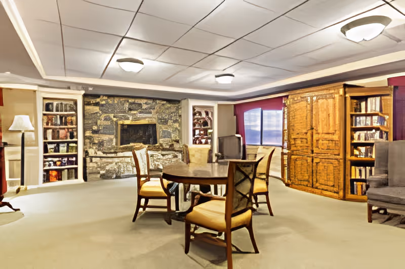 A cozy living room area with a round wooden table surrounded by six cushioned chairs. The room features a stone fireplace in the background flanked by built-in bookshelves filled with books. There is a large wooden cabinet and additional bookshelves on the right side, with a window letting in natural light. The ceiling has recessed lighting fixtures, and the floor is carpeted.
