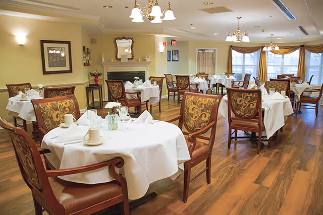 A well-lit dining room with round tables covered in white tablecloths, each set with cups, napkins, and small flower vases. The room features wooden floors, upholstered chairs with patterned backs, a fireplace with a mirror above it, framed artwork on the walls, and large windows with curtains allowing natural light to enter.