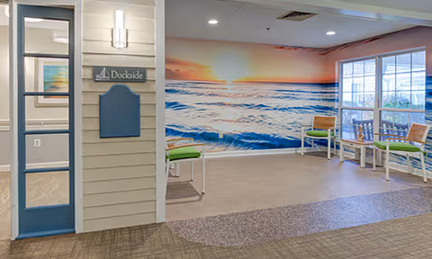 Interior common area with a large wall mural of a sunset over the ocean. The room has several chairs with green cushions and white frames arranged near windows that let in natural light. A sign on the wall reads 'Dockside'.
