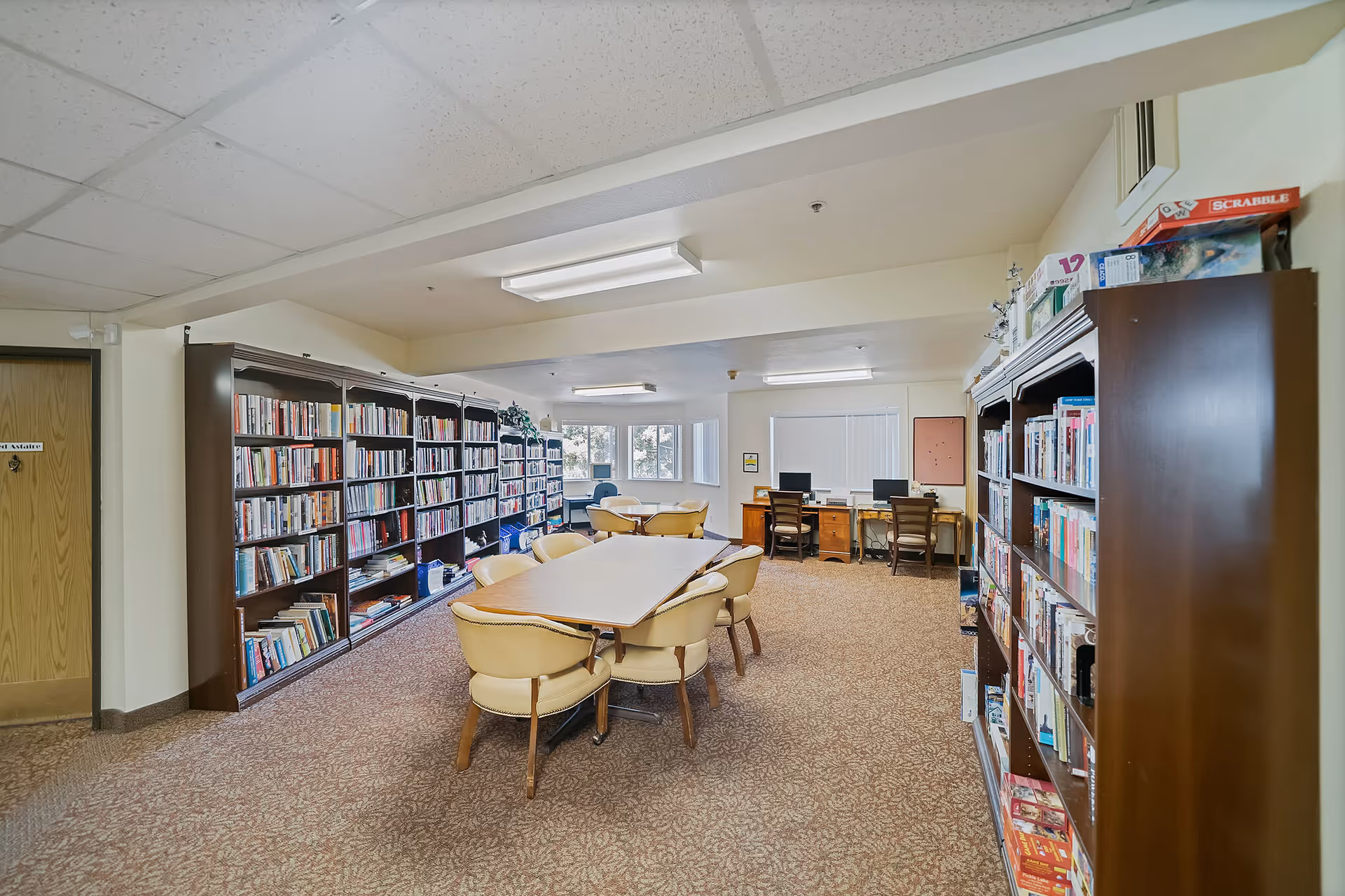 A well-lit room with bookshelves filled with books lining the walls on both sides. In the center, there is a rectangular table surrounded by six beige cushioned chairs. At the far end of the room, there are two desks with computers and chairs in front of windows with blinds. The carpet is patterned, and the ceiling has fluorescent lighting.