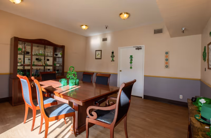 A dining room with a wooden dining table surrounded by six chairs with blue cushions. A wooden china cabinet with glass doors displaying dishes and glassware is against the wall. The room is decorated with green St. Patrick's Day decorations including shamrocks and a leprechaun figure on the door. The walls are painted beige with a purple lower half and a wooden chair rail. The floor is wood, and the room is lit by ceiling lights and sunlight from a window.