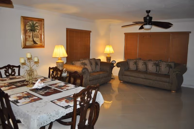 A cozy living and dining area with two brown upholstered sofas adorned with palm tree patterned cushions, a wooden dining table covered with a lace tablecloth and placemats, two table lamps providing warm lighting, wooden blinds covering the windows, a ceiling fan, and a framed palm tree artwork on the wall.