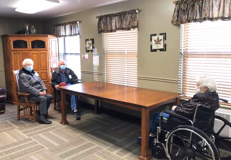 Three elderly residents wearing masks sit around a large wooden table in a communal room, one in a wheelchair.