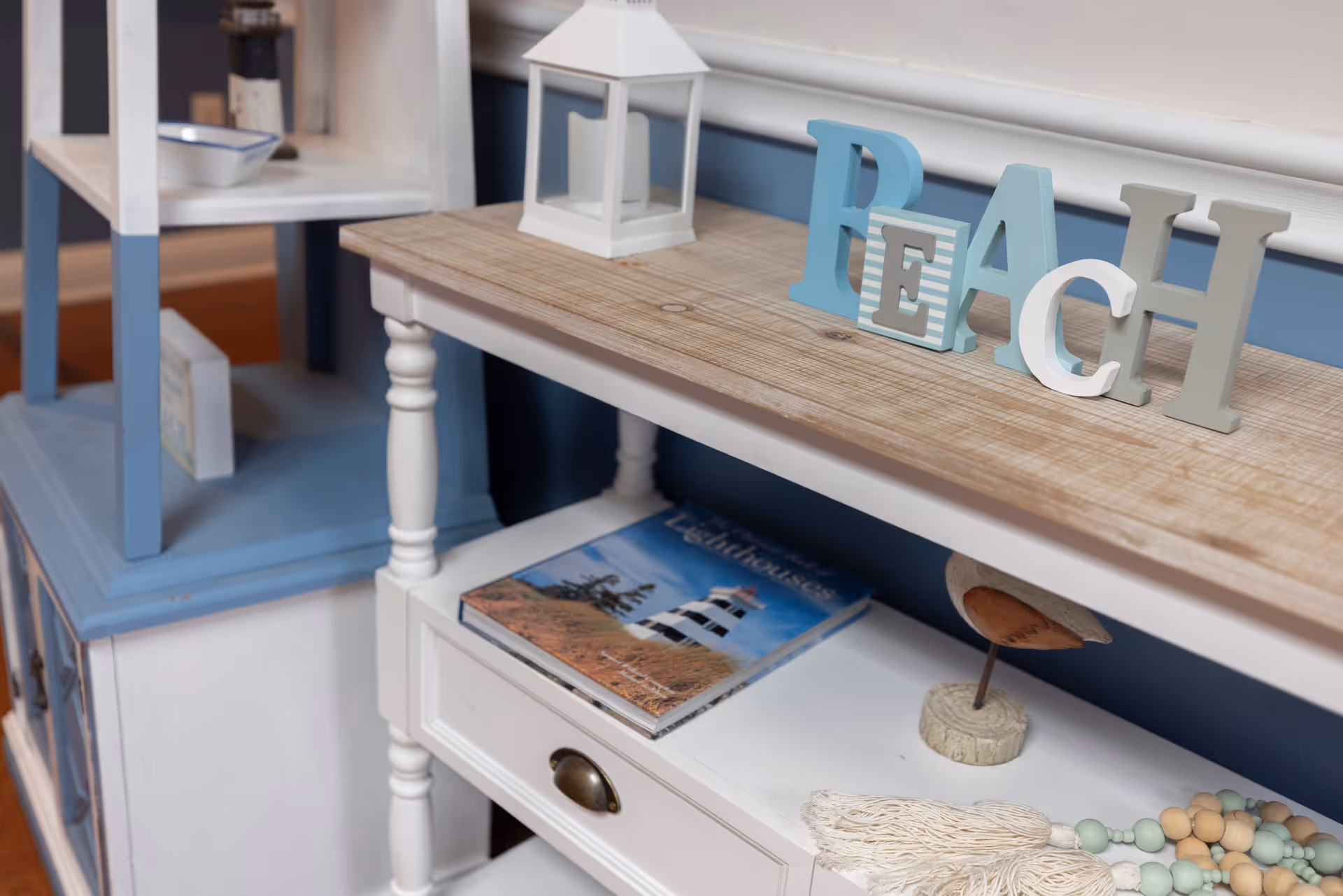 A close-up view of a decorative wooden shelf with a white lantern and wooden letters spelling 'PEACH' on top. Below, there is a book titled 'Lighthouses' and a small decorative item resembling a bird on a stand. The shelf is painted white with a natural wood top, and there is a blue and white cabinet next to it.