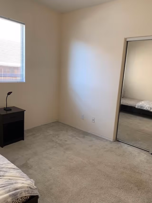 A simple bedroom with beige carpet, cream-colored walls, a window with blinds, a small black nightstand with a lamp, and a mirrored sliding closet door reflecting part of a bed with a blanket.