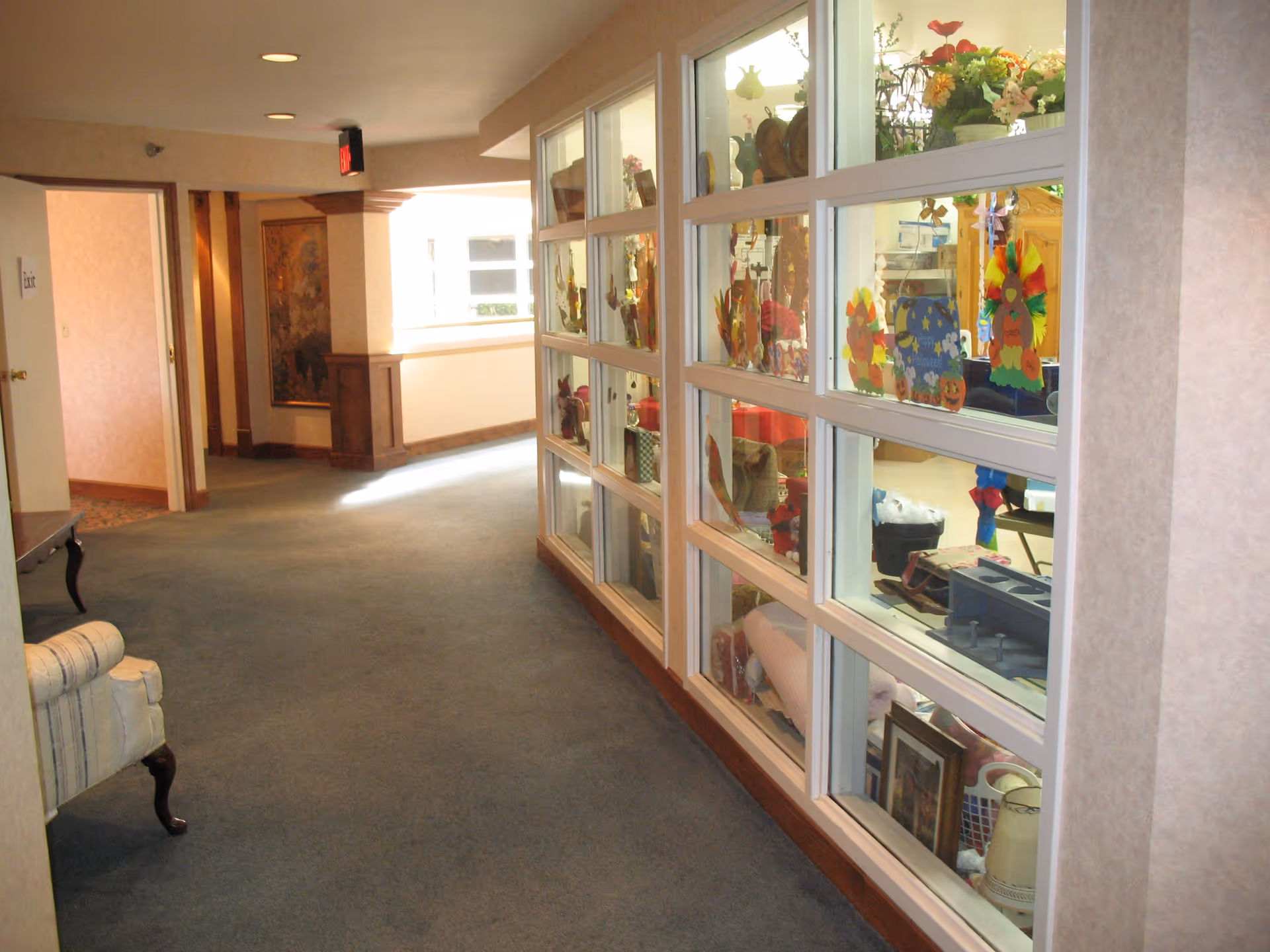 A carpeted hallway in a senior living facility with a glass display case on the right filled with colorful decorations and various items. The hallway has beige walls and a cushioned chair partially visible on the left. There is a painting on the far wall and a door marked 'Exit' at the end of the hallway.
