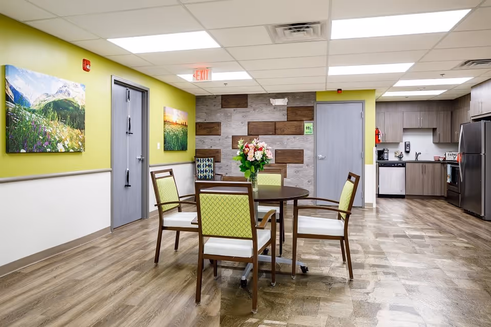 A bright and clean common area in a senior living facility featuring a round table with four chairs around it, a vase with flowers on the table, and a modern kitchen area with stainless steel appliances in the background. The walls are painted light green and white with landscape paintings and a decorative wood panel accent wall.