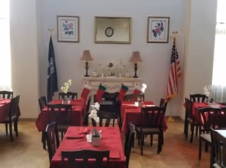 A dining room with several tables covered in red tablecloths, each set with a small white flower centerpiece. At the back of the room is a white mantel decorated with two lamps, framed artwork, and Christmas stockings hanging from it. Two flags, one American and one dark-colored, stand on either side of the mantel. The room is well-lit with natural light coming through windows on both sides.