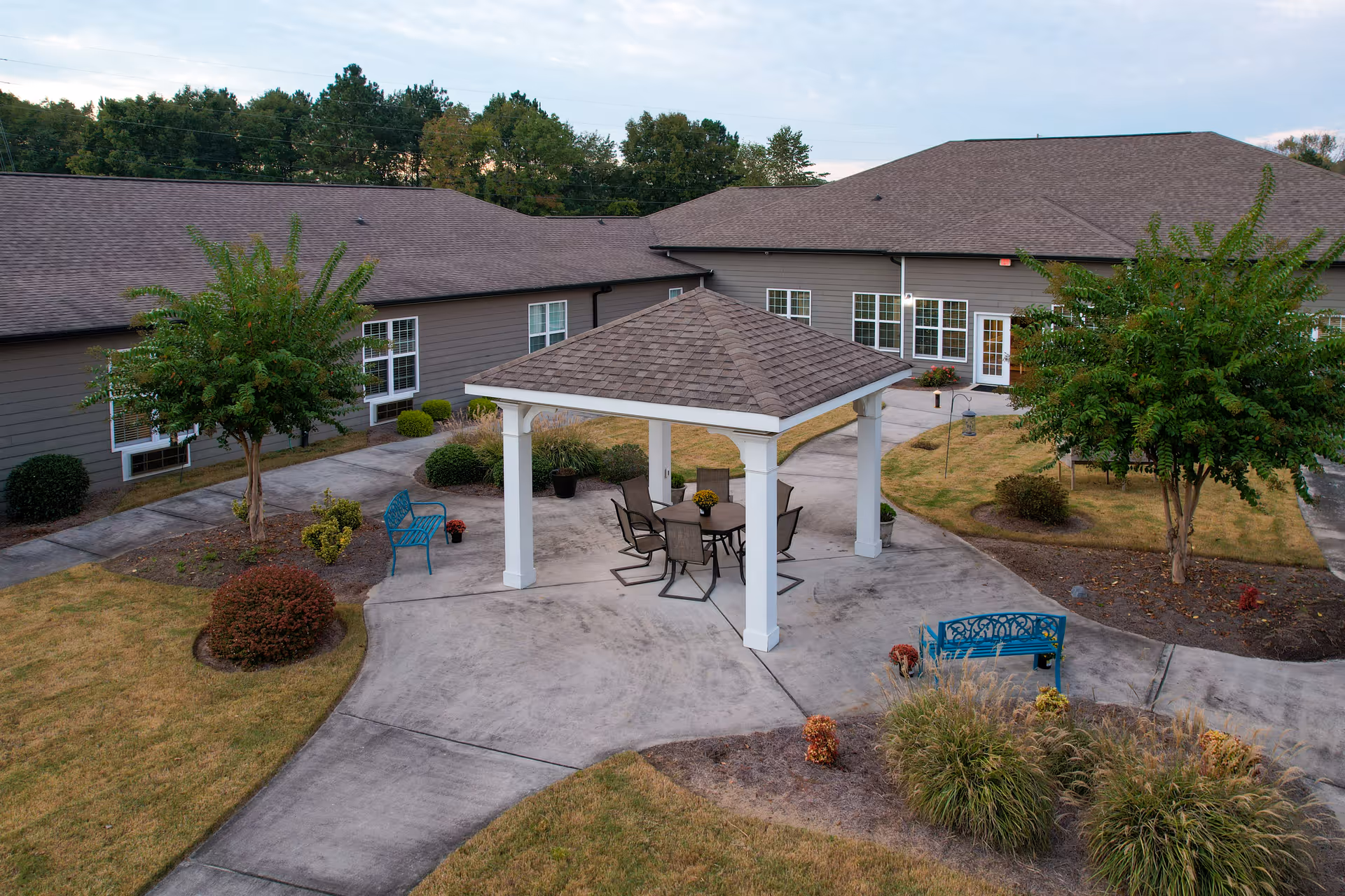 Outdoor courtyard area at Orchard Ridge Residences featuring a covered gazebo with a table and chairs underneath, surrounded by paved walkways, benches, trees, and landscaped bushes with a building in the background.
