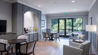 A bright and modern living room area with light wood flooring, gray walls, and large windows letting in natural light. The room features comfortable seating including armchairs and a small sofa, a round dining table with chairs, a built-in cabinet with a coffee station, and a floor lamp. Outside the windows, greenery is visible.