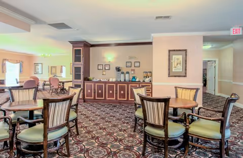 A dining area in a senior living facility with several round wooden tables and cushioned chairs arranged on a patterned carpet. In the background, there is a counter with coffee and beverage dispensers, cups, and framed pictures on the wall. The room is softly lit with wall sconces and ceiling lights, and there is an exit sign visible above a doorway.