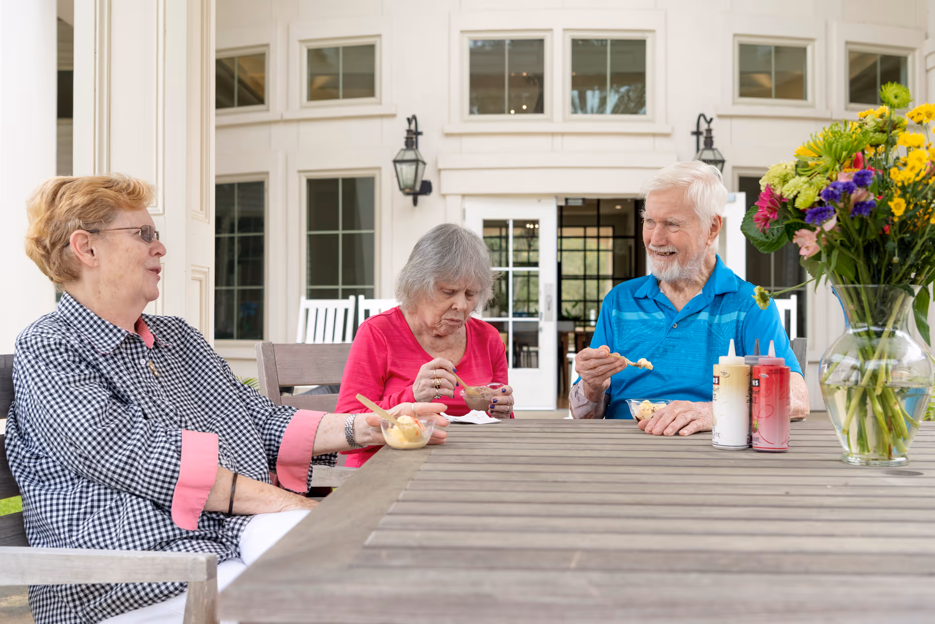 Three elderly people sitting around a wooden outdoor table enjoying ice cream. They are engaged in conversation and smiling. The background shows a building with large windows and white rocking chairs. A vase with colorful flowers and condiment bottles are on the table.