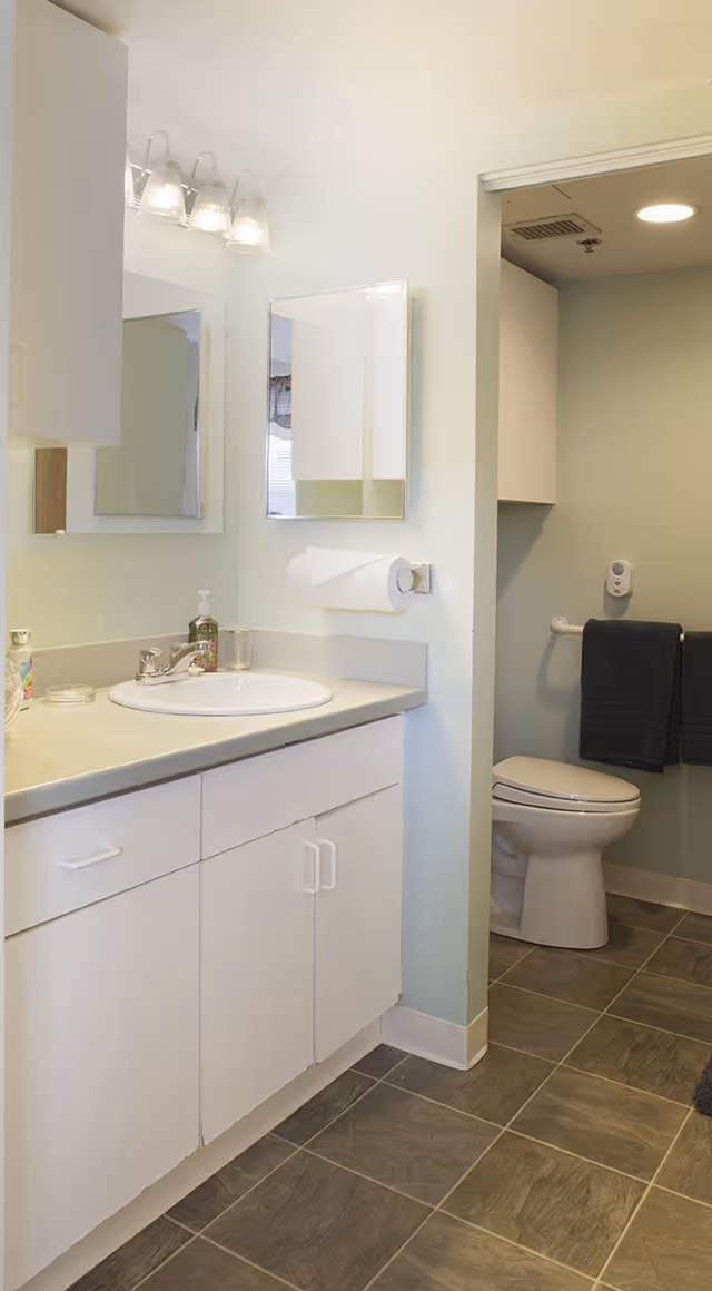 A clean bathroom with a white sink and countertop, white cabinets underneath, a wall-mounted paper towel holder, a mirror above the sink, and a toilet with a dark towel hanging on a grab bar next to it. The floor is tiled with gray tiles.