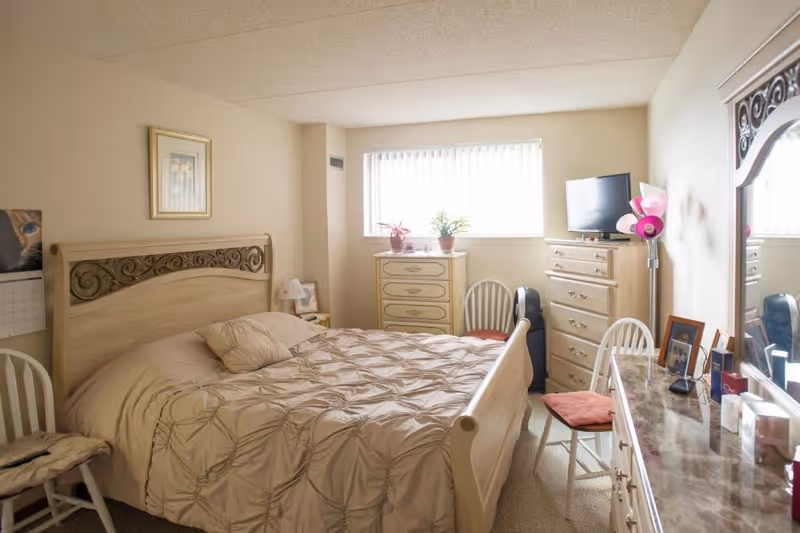 A cozy bedroom with a beige bedspread on a wooden bed frame featuring decorative carvings. The room has a dresser with a TV on top, a chest of drawers, two white chairs with cushions, a floor lamp with pink shades, and a large mirror above a marble-topped dresser. A window with vertical blinds lets in natural light, and there are a few potted plants and framed pictures in the room.