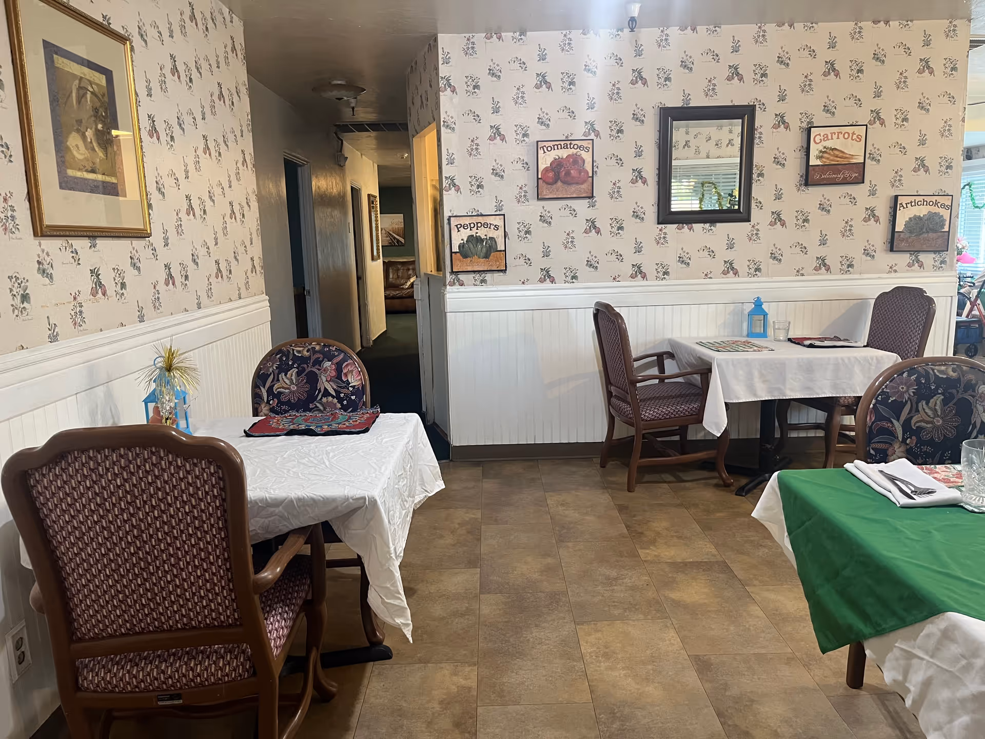 Interior view of a dining area in a senior living facility with tables covered in white and green tablecloths, chairs with patterned upholstery, floral wallpaper, and framed pictures of vegetables on the walls.