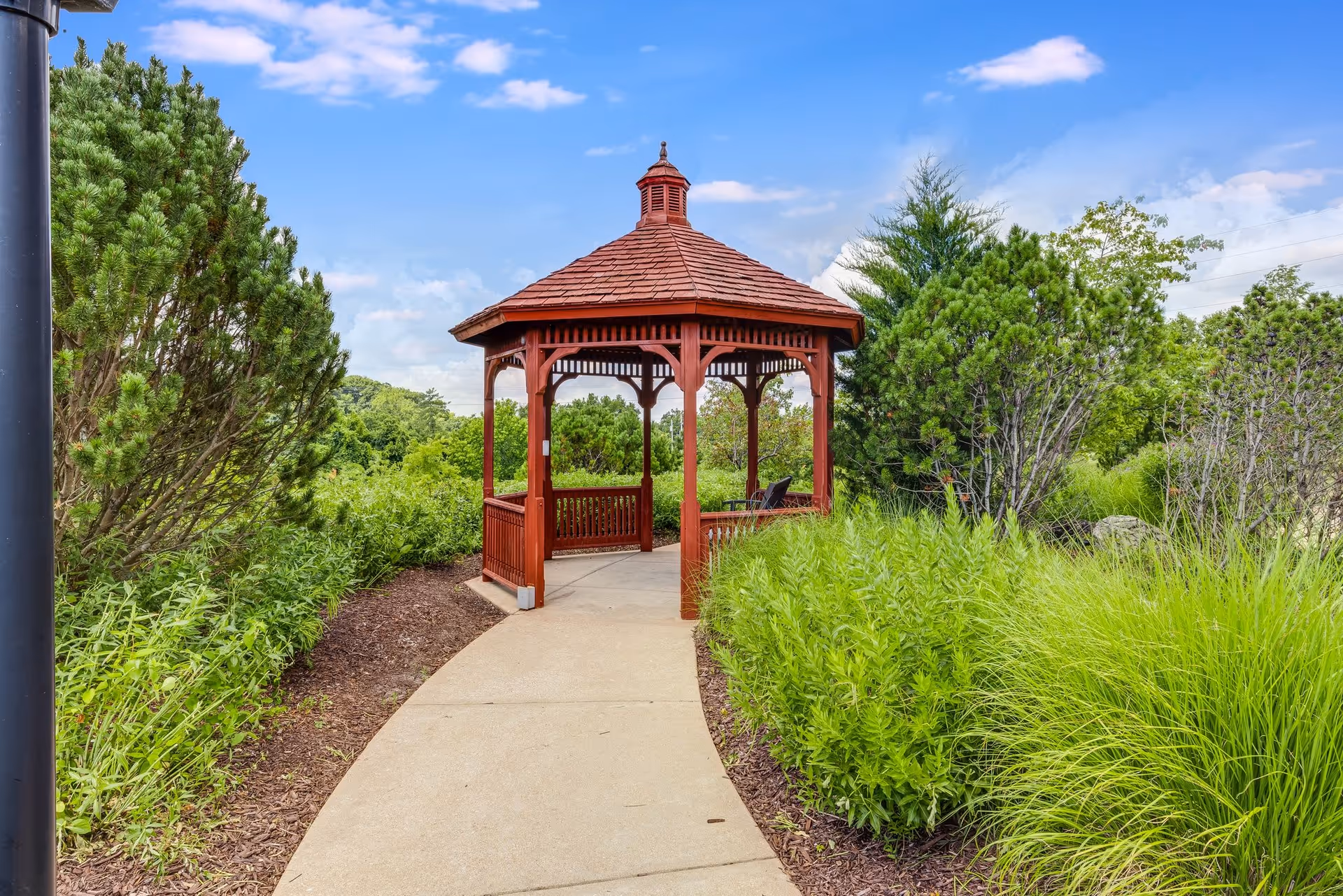 A red wooden gazebo with a shingled roof situated at the end of a curved concrete pathway, surrounded by lush green bushes and trees under a partly cloudy blue sky.