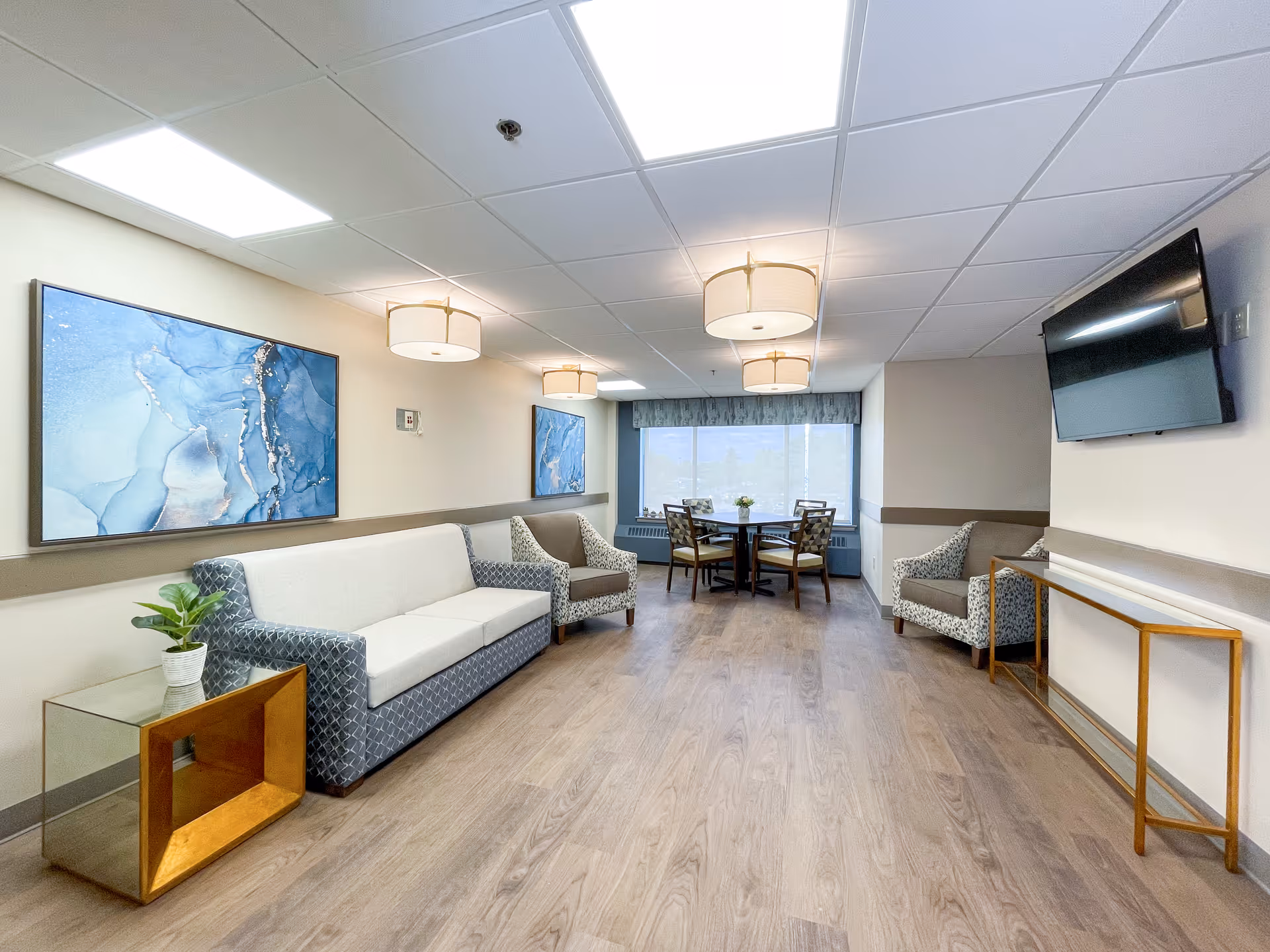 Well-lit communal living area with sofas, armchairs, wall art, a wall-mounted TV, and a small dining table by a window.