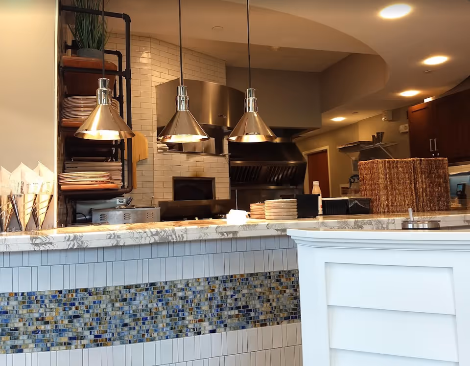 Interior view of a kitchen area with a marble countertop and mosaic tile front. Three metallic pendant lights hang above the counter. Behind the counter, there is a wood-fired oven, a large industrial vent hood, shelves with plates, and various kitchen items.