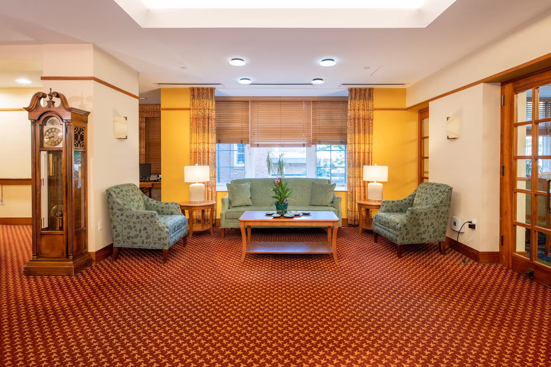 A cozy living room area with a green sofa and two matching armchairs arranged around a wooden coffee table with a potted plant on it. There are two side tables with lamps on either side of the sofa. The room has patterned red carpet, yellow walls with windows covered by blinds and curtains, and a wooden grandfather clock on the left side. A glass door is visible on the right side.