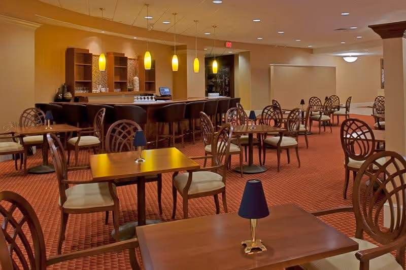 A dining area with multiple wooden tables and chairs arranged on a red carpeted floor. Each table has a small lamp with a dark shade. In the background, there is a bar counter with high chairs and hanging yellow pendant lights above it. The walls are painted beige, and the ceiling has recessed lighting.