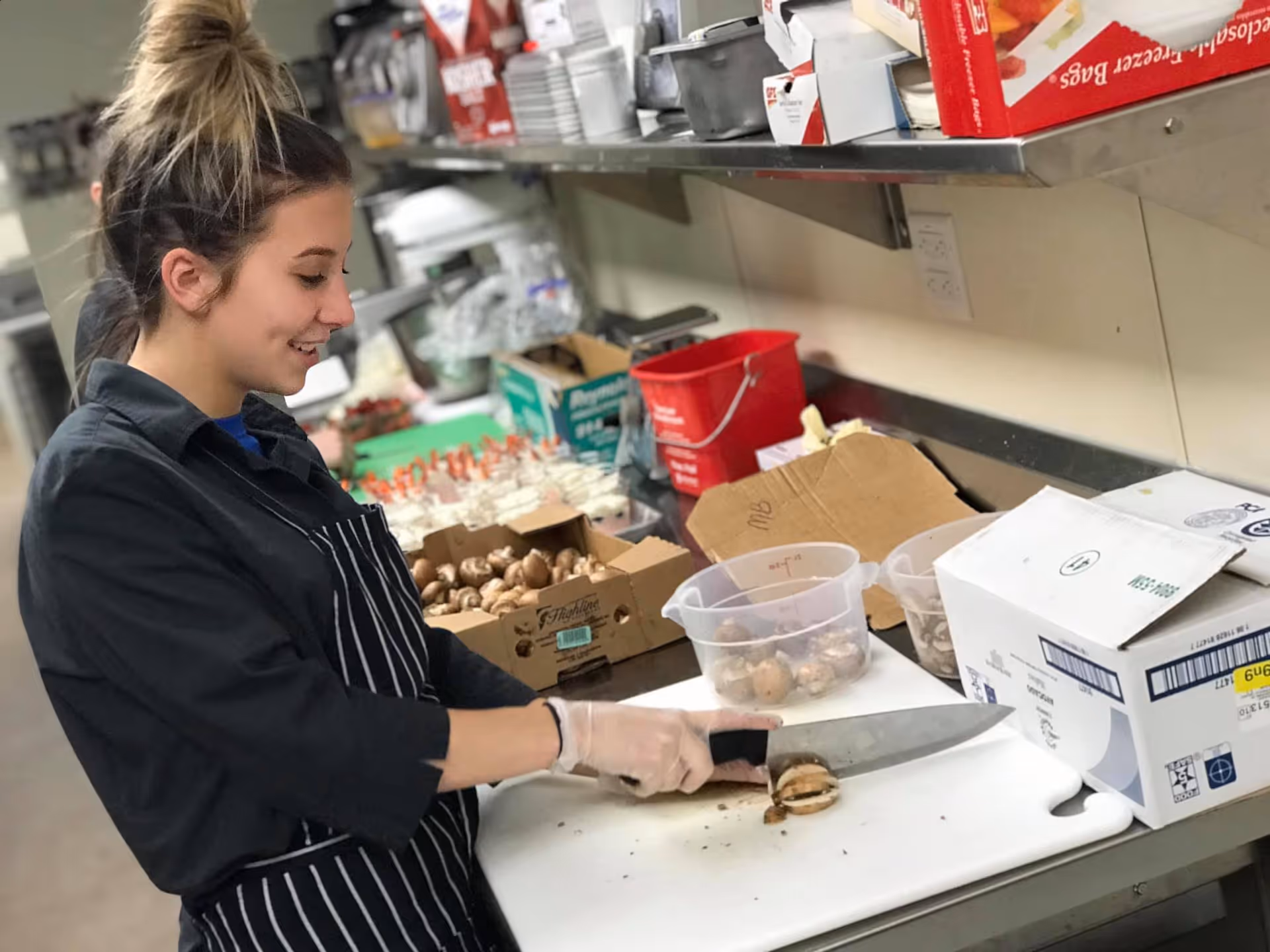 A woman wearing a black shirt and striped apron is slicing mushrooms on a white cutting board in a kitchen. Various kitchen supplies, boxes, and containers with mushrooms are visible on the counter and shelves behind her.
