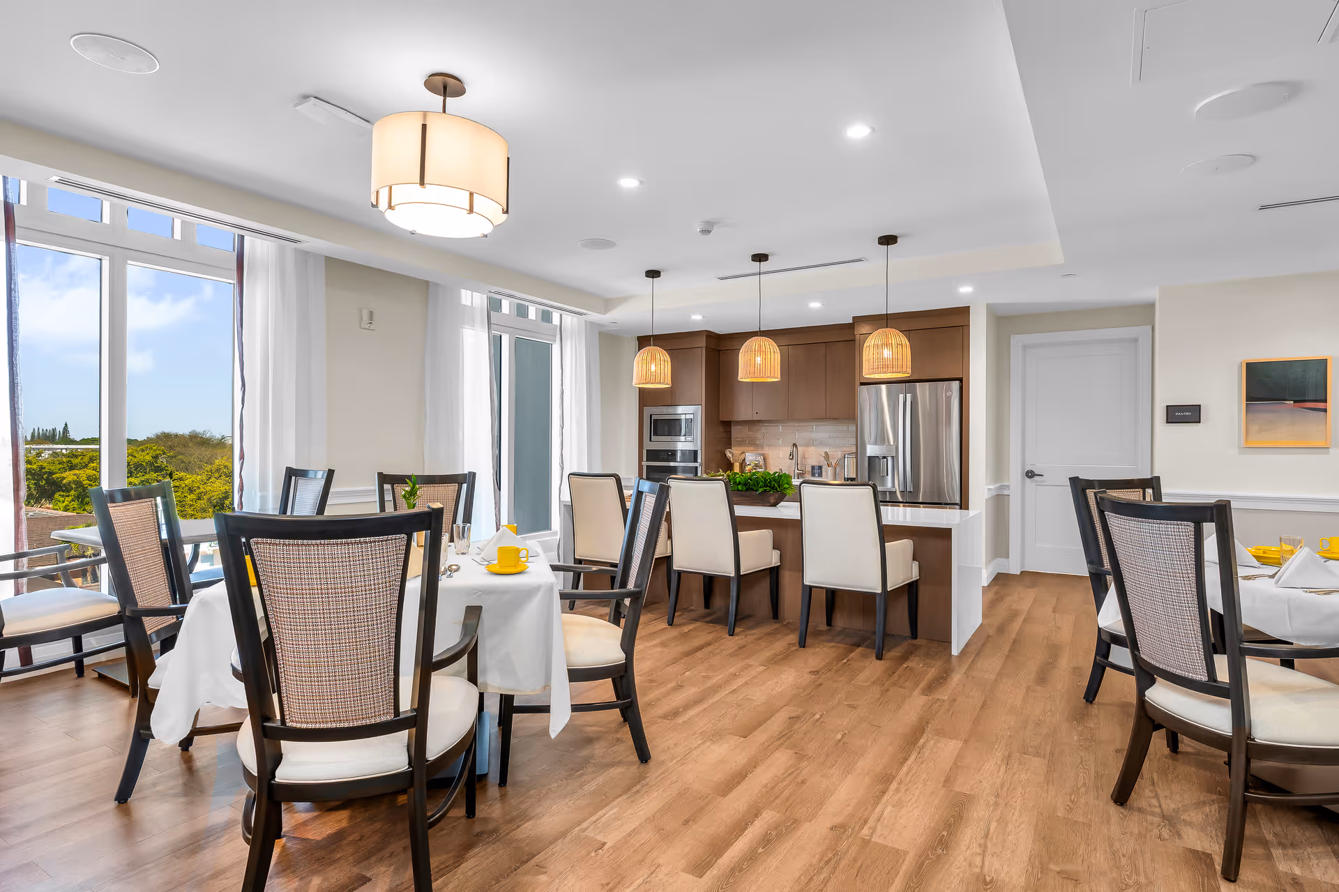 A bright dining area in a senior living facility featuring several round tables covered with white tablecloths, each set with yellow cups and napkins. The room has large windows with white curtains allowing natural light to fill the space. In the background, there is a modern kitchen with wooden cabinets, stainless steel appliances, and three pendant lights hanging over a kitchen island with beige cushioned chairs.