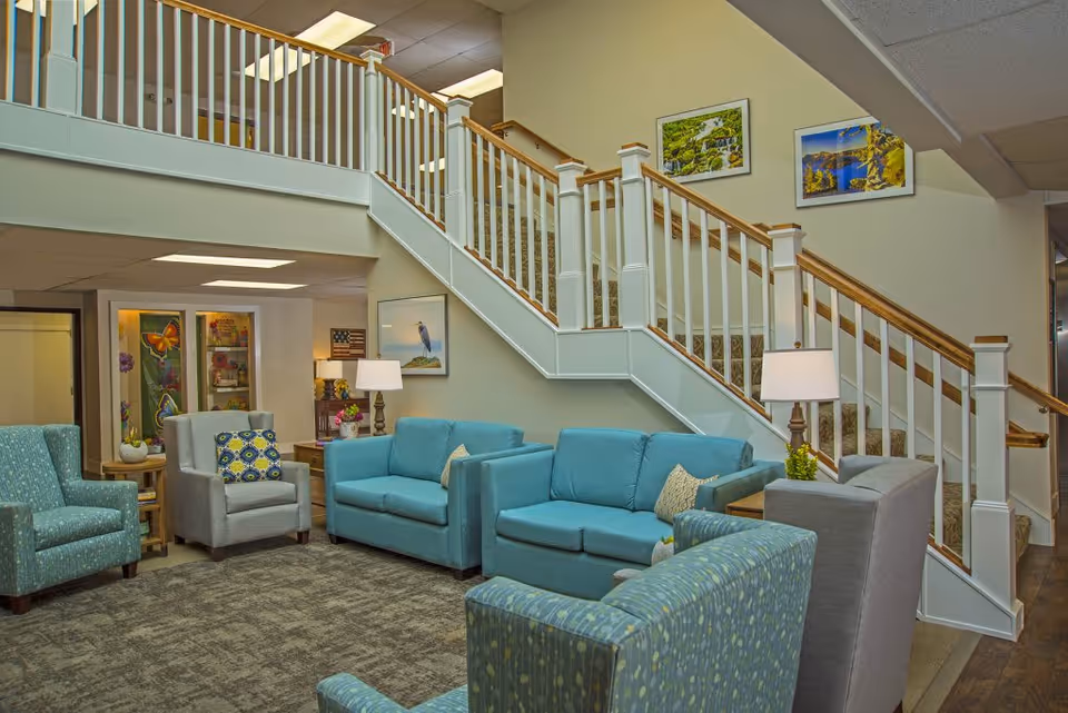 A cozy senior living facility common area with teal and gray upholstered chairs and sofas arranged around a carpeted floor. There are two table lamps on wooden side tables, and a staircase with white railings and wooden handrails leading to an upper floor. The walls are decorated with framed landscape paintings.