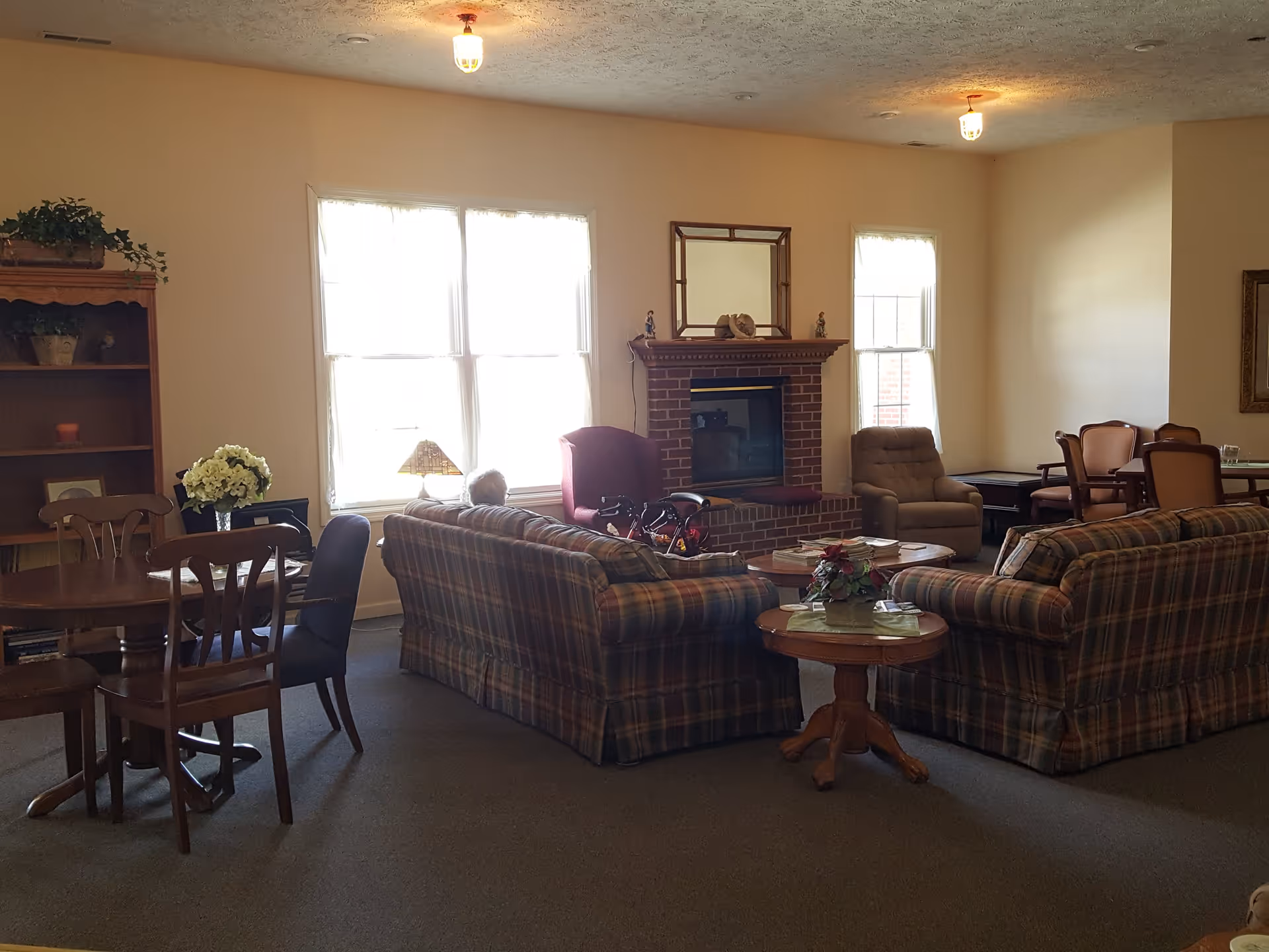 A cozy living room area with two plaid sofas facing a brick fireplace. There is a small round wooden table with a flower arrangement in front of the sofas. To the left, there is a wooden dining table with chairs and a bookshelf with plants and decorative items. The room is softly lit by ceiling lights and natural light coming through the windows.