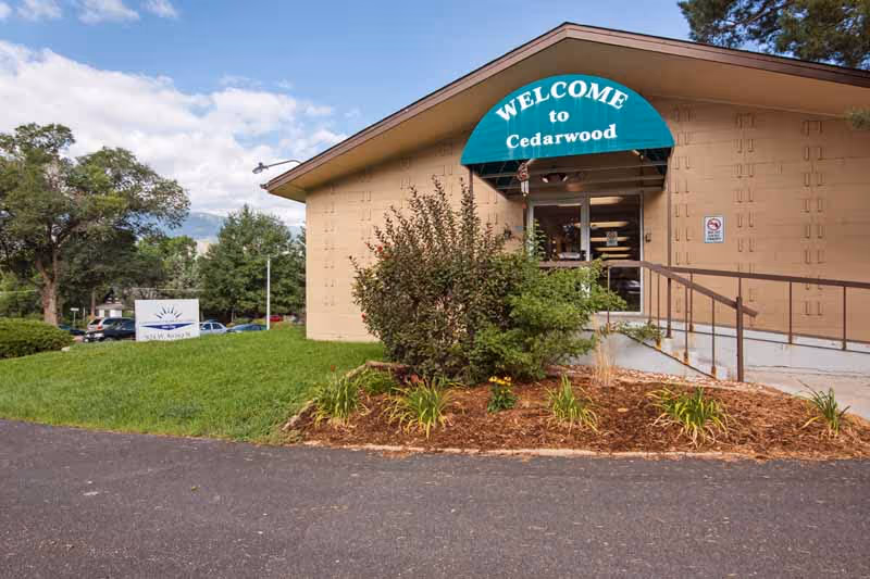 Exterior view of a single-story building with a green awning that reads 'WELCOME to Cedarwood' above the entrance door. There is a small landscaped area with bushes and plants in front of the building, a paved driveway, and trees in the background under a partly cloudy sky.