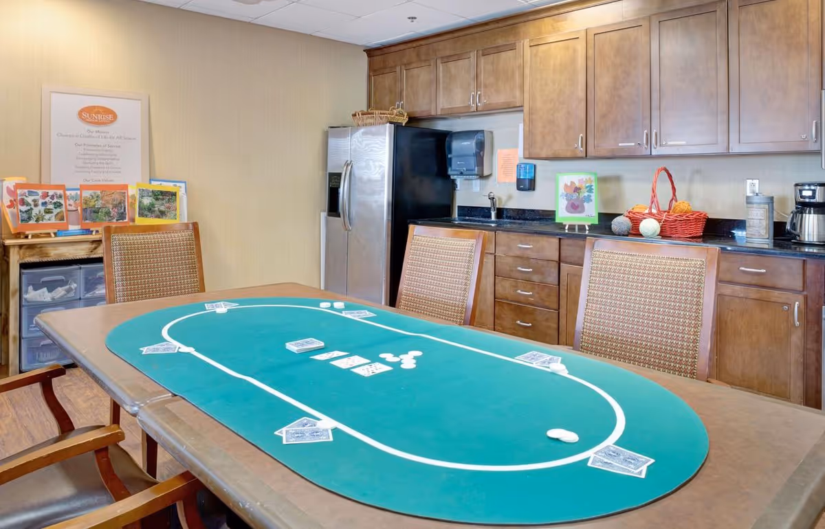Communal activity room with a green felt poker table in the foreground, chairs around it, and a kitchenette with cabinets and refrigerator in the background.