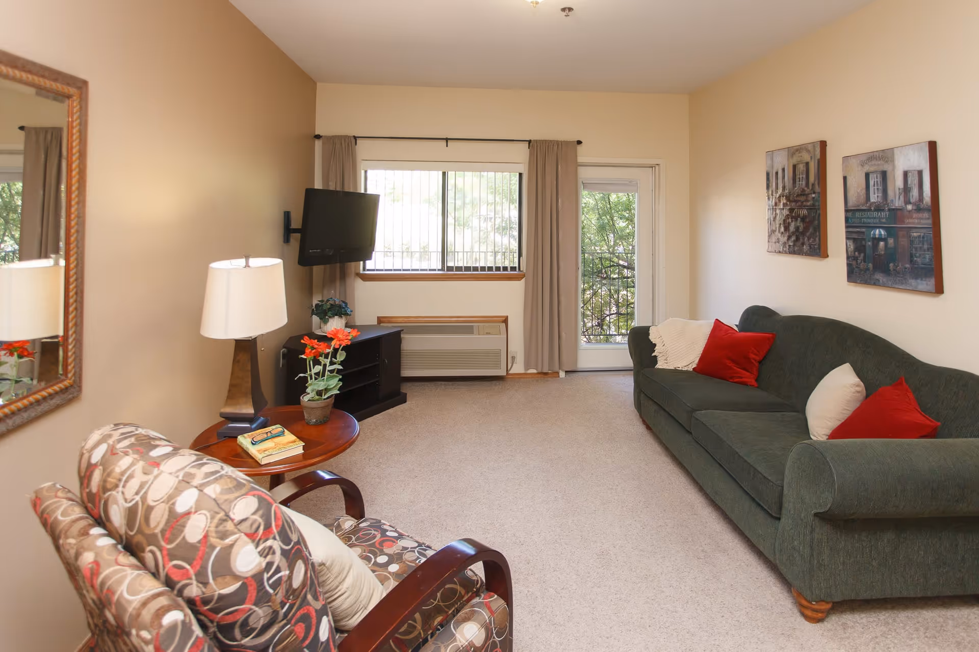 A cozy living room with a green sofa adorned with red and white pillows, a patterned armchair, a wooden side table with a lamp, a book, and a pair of glasses. There is a wall-mounted TV near a window with beige curtains and a door leading outside. Two framed paintings hang on the wall above the sofa.