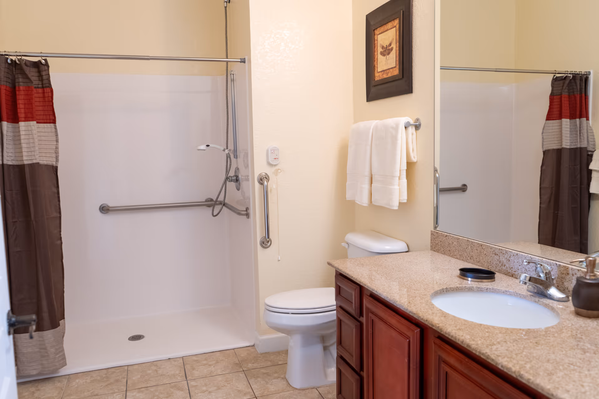 A clean bathroom featuring a walk-in shower with a grab bar and a shower curtain in brown, red, and beige stripes. There is a toilet next to the shower, a towel rack with two white towels, a framed picture above the towel rack, and a granite countertop with a sink and faucet. The floor is tiled and a large mirror is mounted above the countertop.