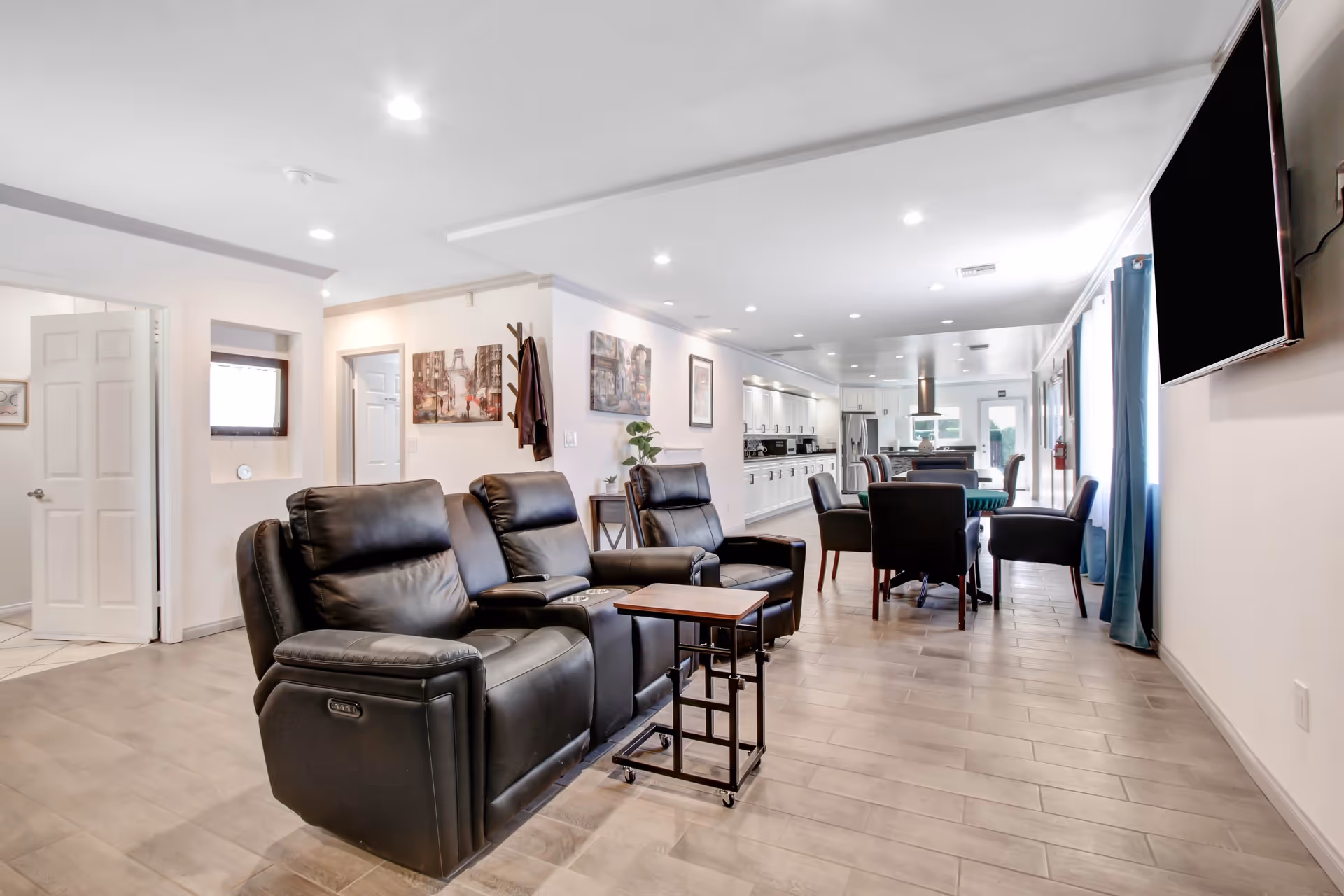 A spacious, well-lit common area in a senior living facility featuring black leather recliners with a small wooden side table on wheels, a dining table with chairs, white cabinetry along the far wall, and a large flat-screen TV mounted on the right wall. The room has light-colored walls, wood-look tile flooring, and several pieces of artwork and a coat rack on the walls.