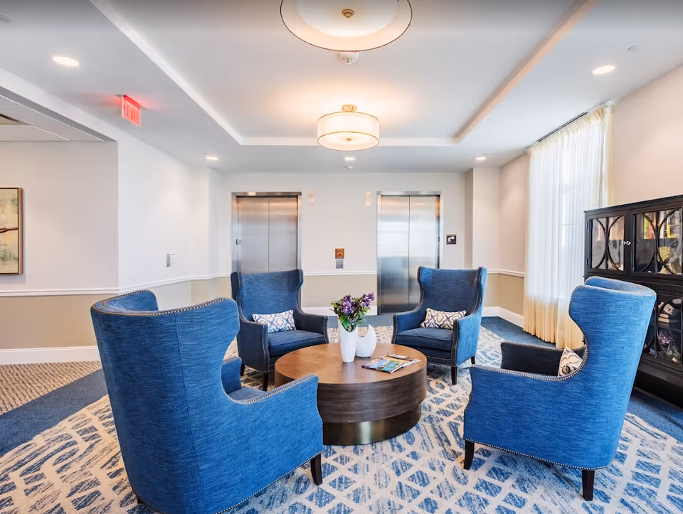 A bright and modern seating area with four blue upholstered armchairs arranged around a round wooden coffee table with a vase of purple flowers and magazines. The room has a patterned carpet, two stainless steel elevator doors on the back wall, a large window with sheer curtains on the right, and a dark wooden cabinet with glass doors. Ceiling lights illuminate the space.