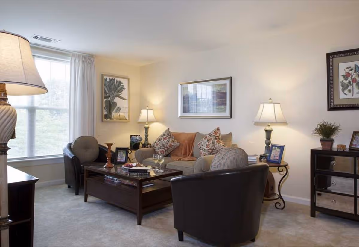 A cozy living room with a beige sofa adorned with patterned cushions and a throw blanket, two dark armchairs, a wooden coffee table with books and decorative items, two table lamps on side tables, framed artwork on the walls, and a large window with white curtains letting in natural light.