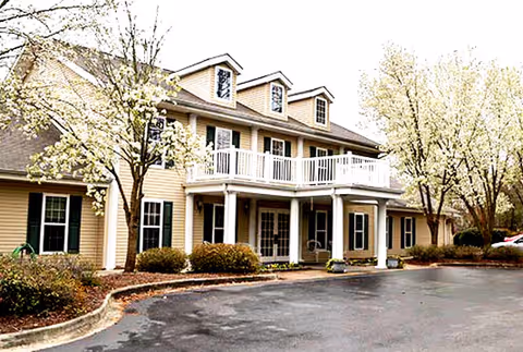 Exterior view of a two-story beige building with white trim and a balcony above the entrance, surrounded by trees with light-colored blossoms and a paved driveway in front.