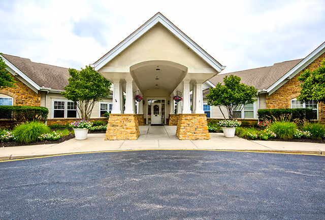 Front entrance of a senior living facility with a covered driveway supported by white columns and stone bases. The building has a combination of stone and siding exterior with windows and landscaped greenery including trees and flower pots on either side of the entrance.