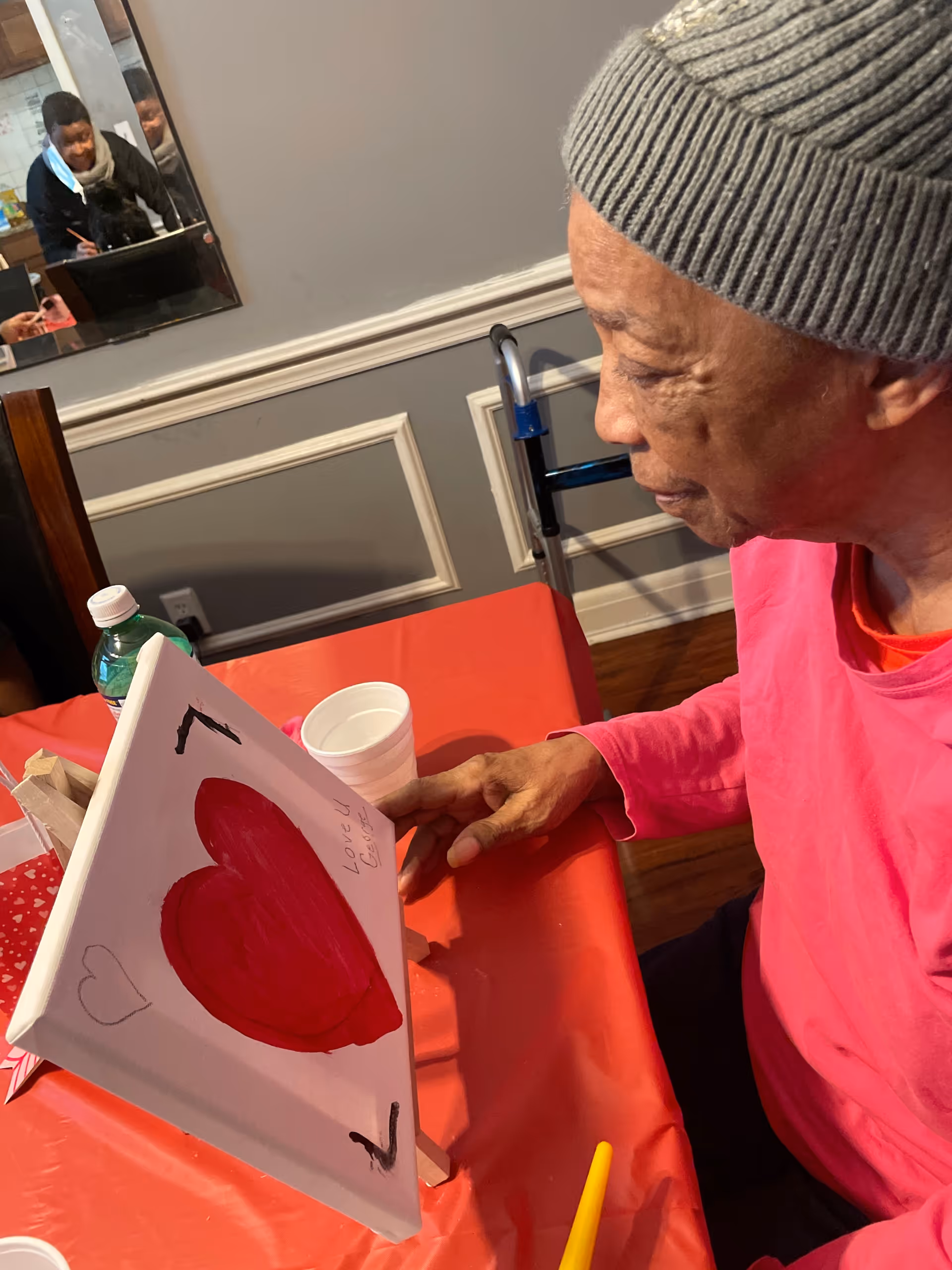 An elderly person wearing a gray knit hat and pink shirt is sitting at a table covered with a red tablecloth, looking at a canvas with a large red heart painted on it. The canvas is propped up on a small easel. In the background, a walker is visible against the wall, and a mirror reflects another person in the room.