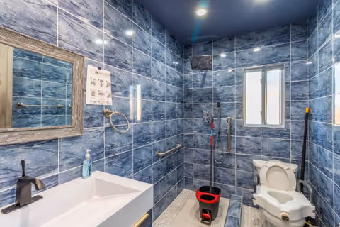 Accessible bathroom with blue tiled walls, a sink and mirror on the left, grab bars and shower area in the center, and a raised toilet with medical seat on the right.