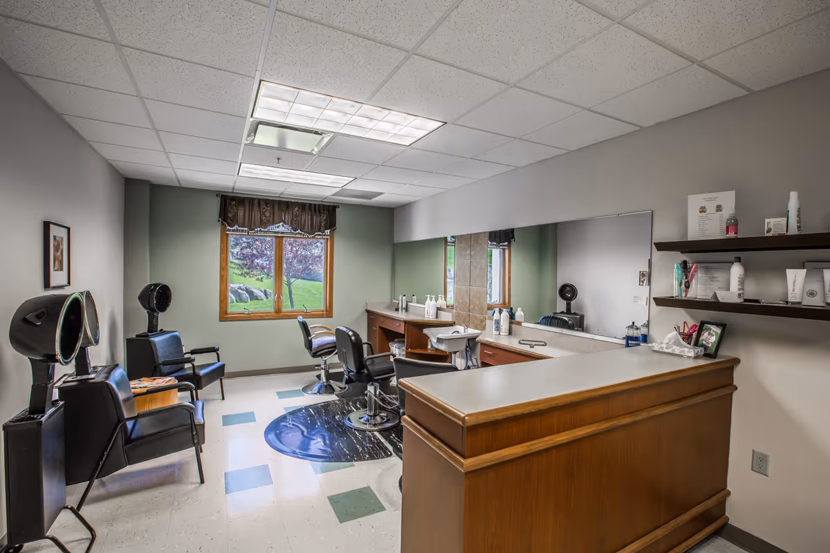 Interior of a hair salon room with two black salon chairs, hair dryers, a large mirror on the wall, a wooden reception counter, shelves with hair products, and a window showing green grass and trees outside.