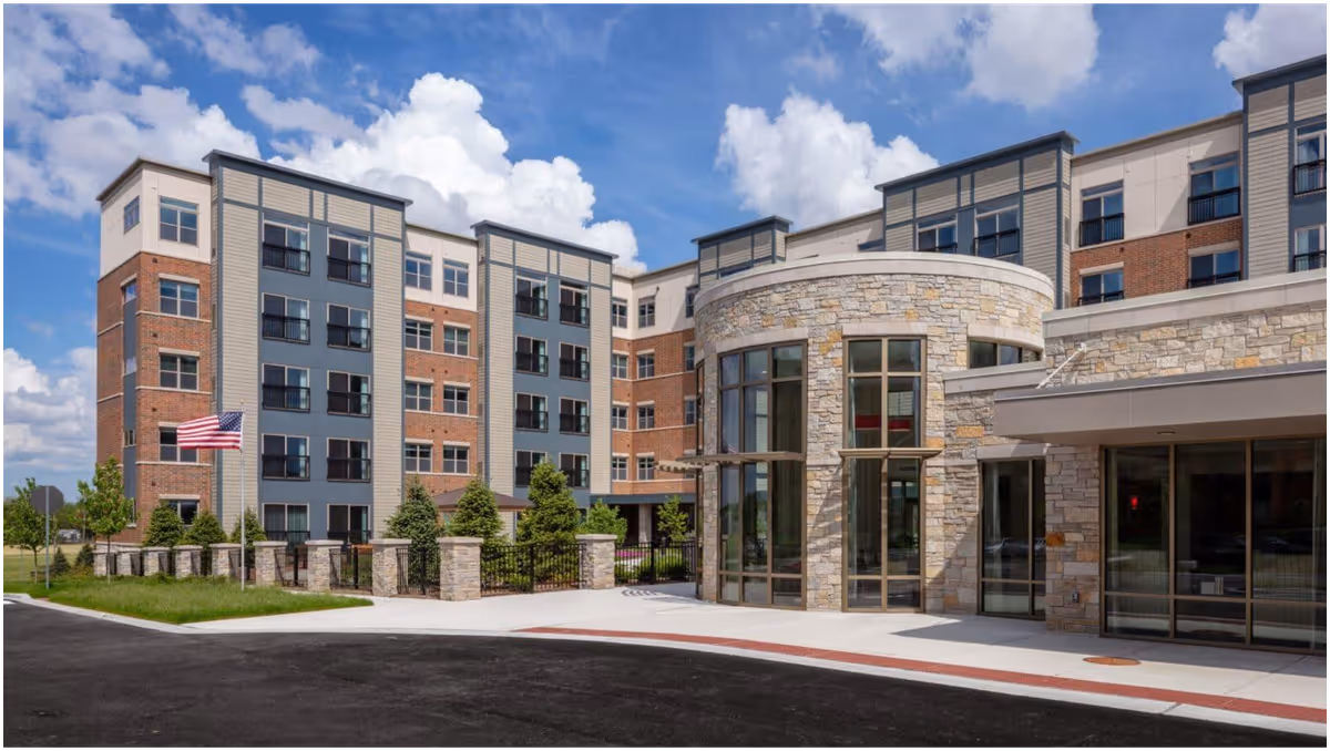 Exterior view of a multi-story senior living facility building with a combination of brick and stone facade, large windows, and an American flag on a flagpole. The entrance features a rounded stone structure with tall windows and a covered walkway.