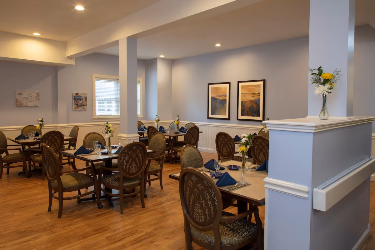Dining room with several neatly set tables, upholstered chairs, floral centerpieces, and framed artwork on soft blue walls.