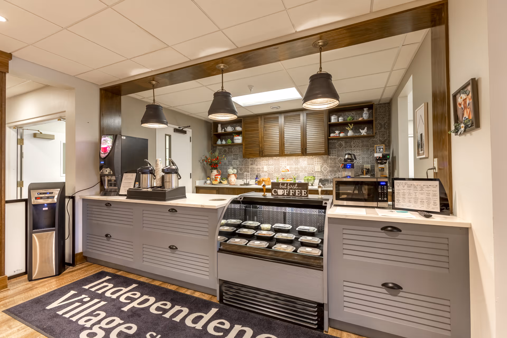 Interior view of a coffee and snack station area in a senior living facility. The space features a counter with a refrigerated display case containing packaged food items, a microwave, coffee dispensers, and decorative shelves with various items. Three pendant lights hang from the ceiling above the counter. A water cooler is visible to the left, and a large rug on the floor reads 'Independence Village'.