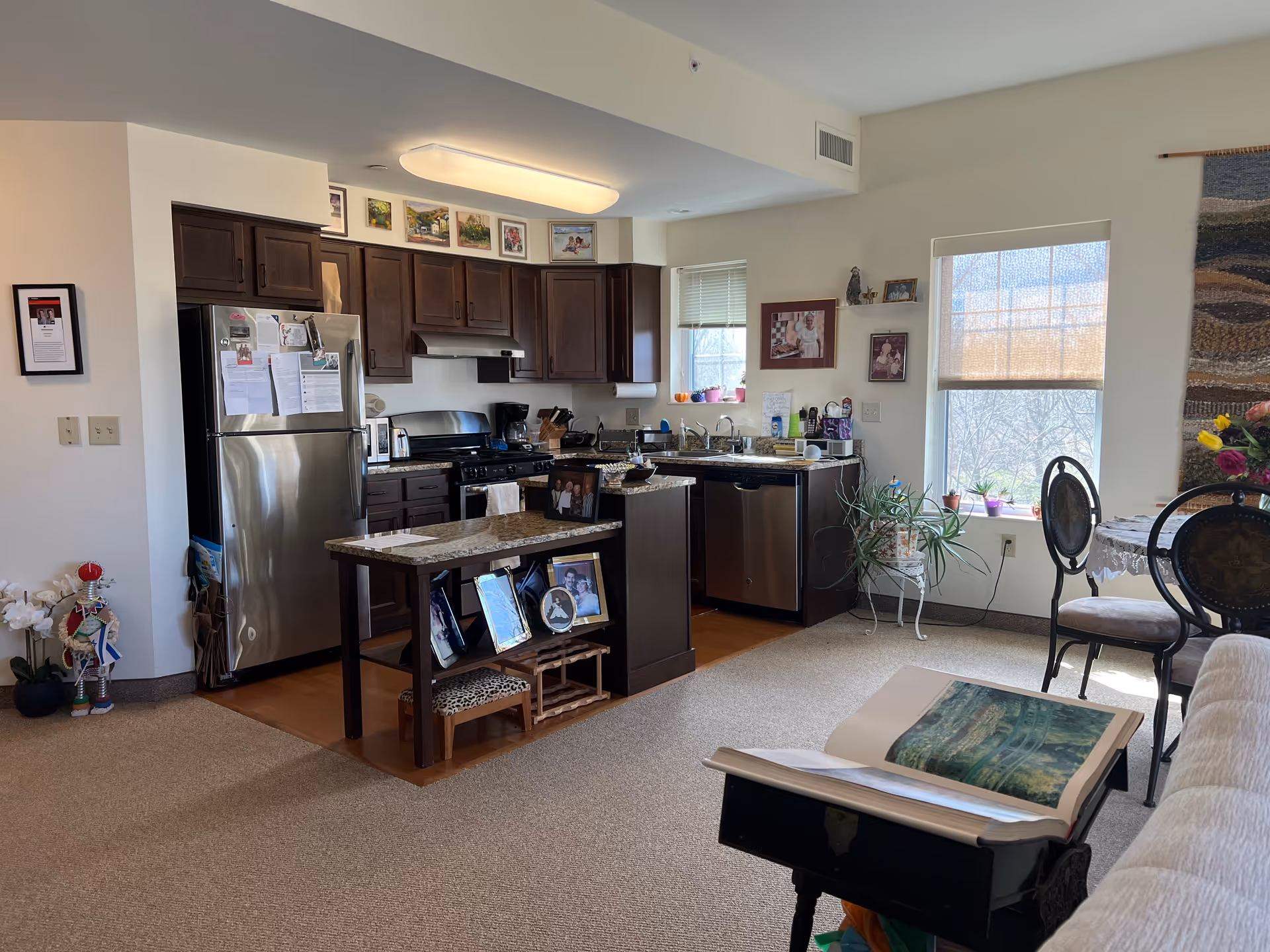 Interior view of a cozy kitchen and dining area in a senior living facility. The kitchen features dark wood cabinets, stainless steel appliances including a refrigerator, stove, and dishwasher, and a small island with framed photos on the shelves. The dining area has a round table with chairs, a window with a shade, and a plant on a stand. The room is well-lit with natural light coming through the windows.