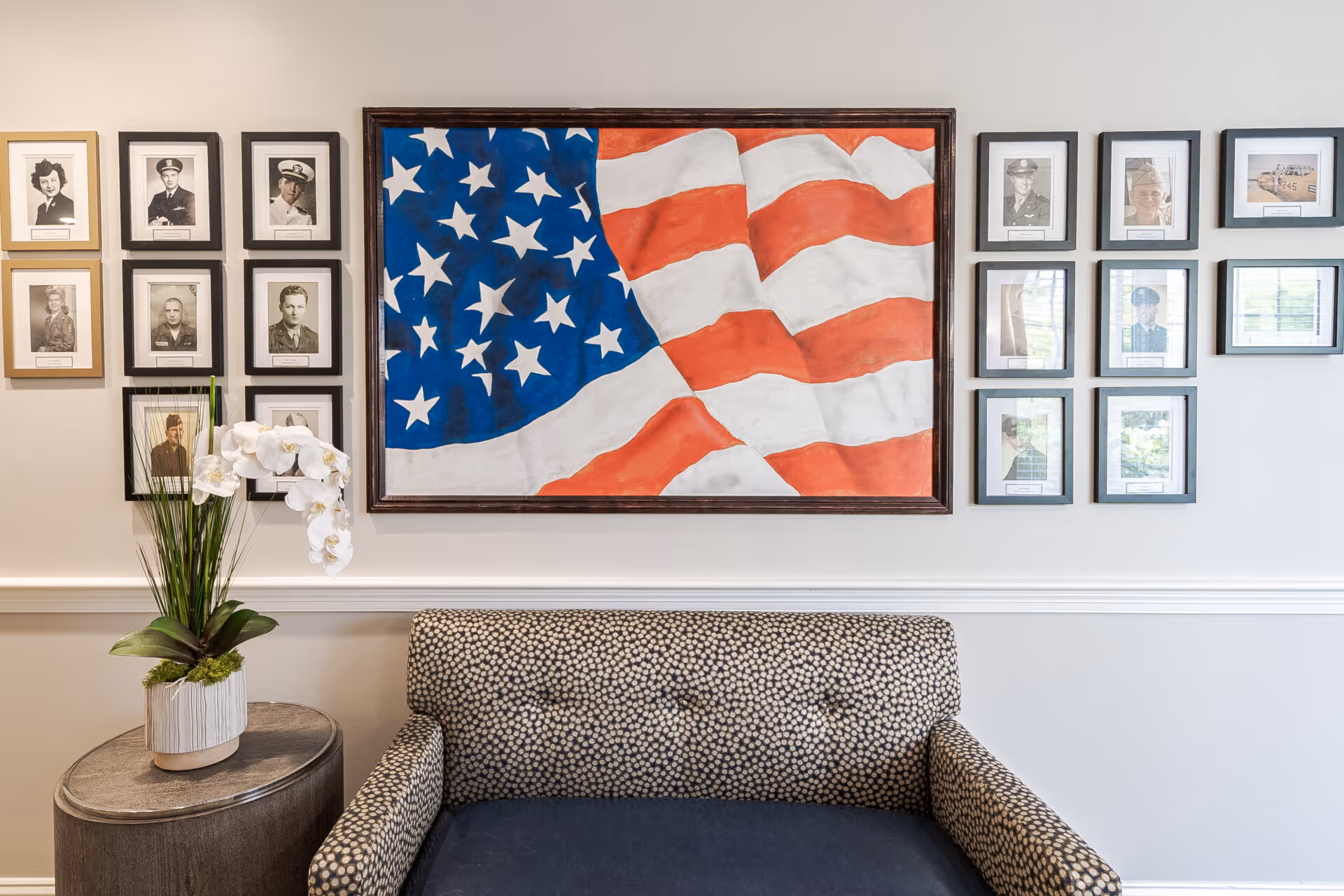 A sitting area with a patterned armchair and a small round side table holding a white orchid plant. Above the chair is a large framed painting of an American flag, flanked by multiple framed black-and-white photographs of military personnel on both sides.