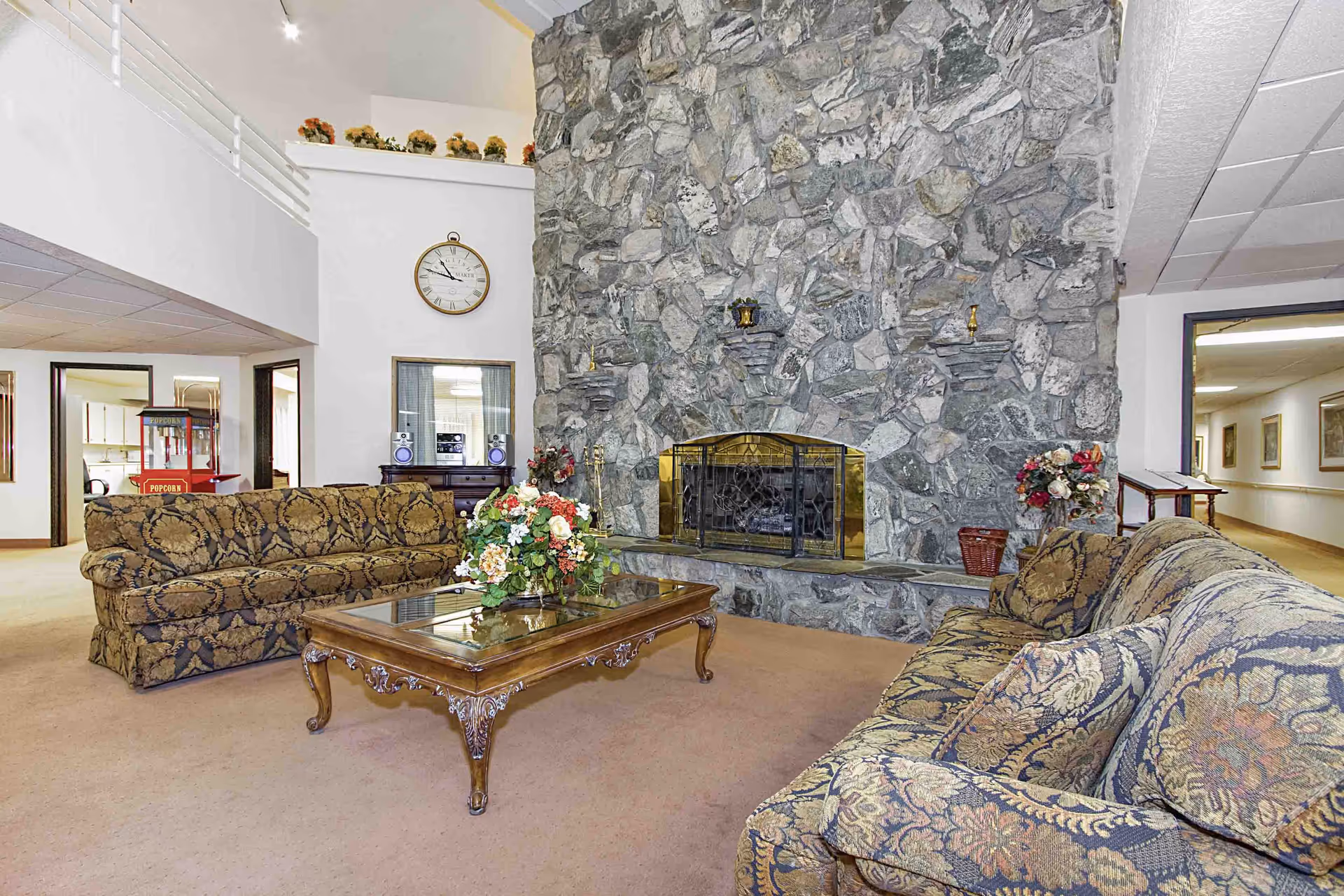 A cozy living room area in a senior living facility with two patterned sofas facing a large stone fireplace. A wooden coffee table with a floral arrangement is centered between the sofas. The room has a high ceiling with a clock on the wall and decorative flowers on a ledge above. There is a popcorn machine visible in the background and a hallway leading to other rooms.