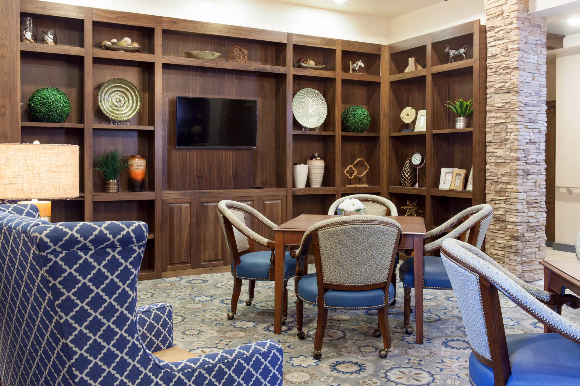 A cozy common area with a wooden table surrounded by six upholstered chairs, a blue patterned armchair, and built-in wooden shelves displaying decorative items and a flat-screen TV mounted on the wall. The room features a stone pillar and a patterned carpet.